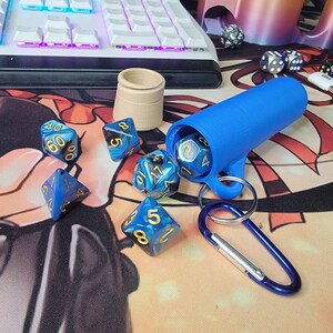 Puede incluir: Una bolsa de dados azul con un mosquetón, que contiene seis dados azules y dorados con números negros. Los dados están esparcidos sobre una superficie con un patrón negro y azul.