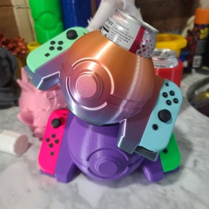 May include: A 3D printed Pokeball-themed Nintendo Switch controller holder with a purple, blue, and orange color scheme. The holder has a slot for a can of soda and can hold two controllers.
