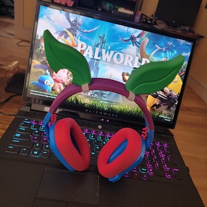 Puede incluir: Un auricular para juegos rojo y azul con cubiertas de oído en forma de hojas verdes. El auricular está sentado en una computadora portátil negra con un teclado colorido. La pantalla de la computadora portátil muestra el juego "Palworld" con un fondo azul y verde.