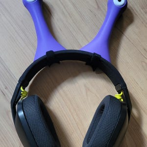May include: A purple headset stand shaped like a snail with two eye stalks. The stand holds a black headset with yellow accents. The earcups are black and padded. The stand is on a wooden surface.