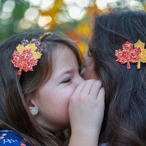 May include: A young girl and an adult with matching fall-themed hair accessories. The girl wears a headband with a glittery red, yellow, and orange leaf. The adult has a similar leaf-shaped hair clip. The background is blurred.