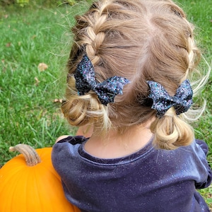 May include: Two black glittery bat-shaped hair bows attached to braided hair. The bows feature a textured, sparkling finish and are shaped like bat wings, secured on each side of the head to hold the braided sections in place.