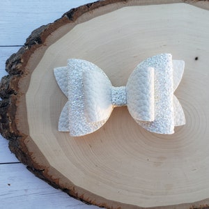 Puede incluir: Un lazo para el pelo blanco brillante con una gran forma de lazo en capas. El lazo est&aacute; sobre una superficie de madera marr&oacute;n claro.