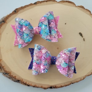 May include: Two sparkly hair bows with a mermaid scale design. One bow is pink and blue, the other is purple and pink.