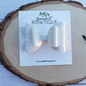 Puede incluir: Un lazo blanco con purpurina y acabado texturizado. El lazo se muestra en una tarjeta blanca con el texto "lynnlett BOWTIQUE" y una direcci&oacute;n de sitio web.