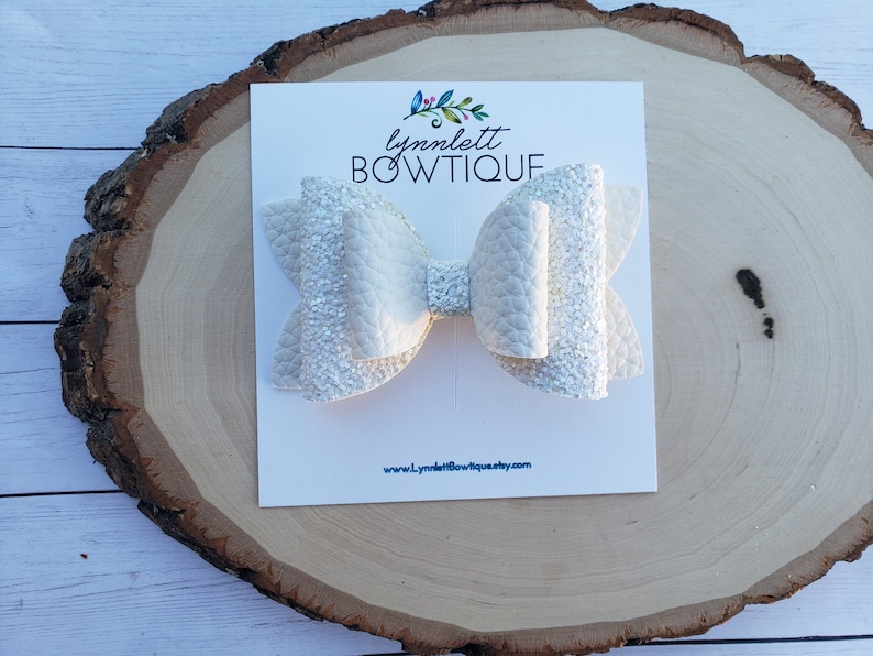 Puede incluir: Un lazo blanco con purpurina y acabado texturizado. El lazo se muestra en una tarjeta blanca con el texto "lynnlett BOWTIQUE" y una direcci&oacute;n de sitio web.