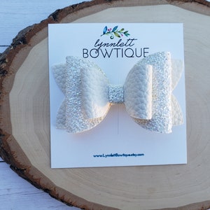 Puede incluir: Lazo blanco con purpurina y acabado texturizado. El lazo se muestra en una tarjeta blanca con el texto "lynnlett BOWTIQUE" y "www.lynnlettbowtique.etsy.com".