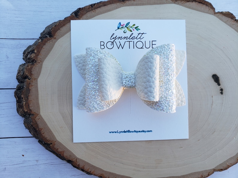 Puede incluir: Lazo blanco con purpurina y acabado texturizado. El lazo se muestra en una tarjeta blanca con el texto "lynnlett BOWTIQUE" y "www.lynnlettbowtique.etsy.com".