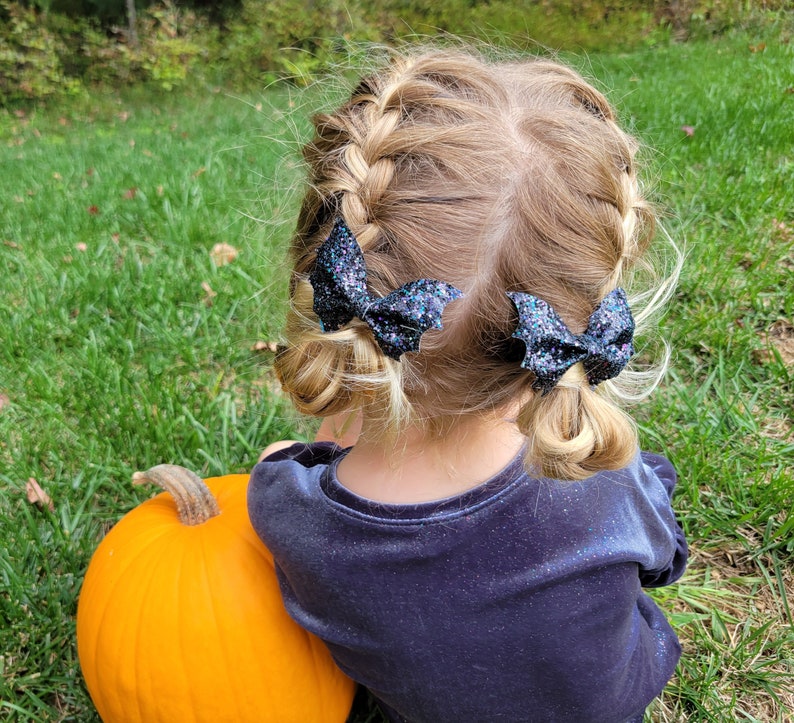 May include: Two black glittery bat-shaped hair bows attached to braided hair. The bows feature a textured, sparkling finish and are shaped like bat wings, secured on each side of the head to hold the braided sections in place.