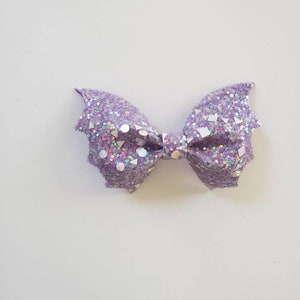 May include: A bow-shaped hair accessory featuring a light purple glitter finish with embedded iridescent geometric shapes including circles, triangles, and squares. The edges of the bow are pointed, resembling bat wings, with a central knot detail.