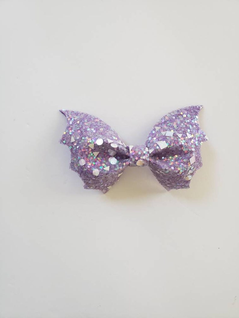 May include: A bow-shaped hair accessory featuring a light purple glitter finish with embedded iridescent geometric shapes including circles, triangles, and squares. The edges of the bow are pointed, resembling bat wings, with a central knot detail.