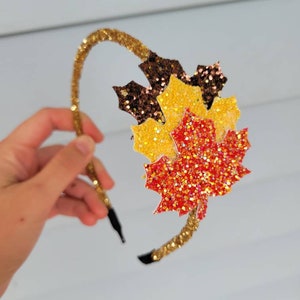 May include: A gold glitter headband with a cluster of glittery maple leaves in shades of brown, yellow, and red. The headband is held by a hand against a light background. The leaves are layered to create a 3D effect.