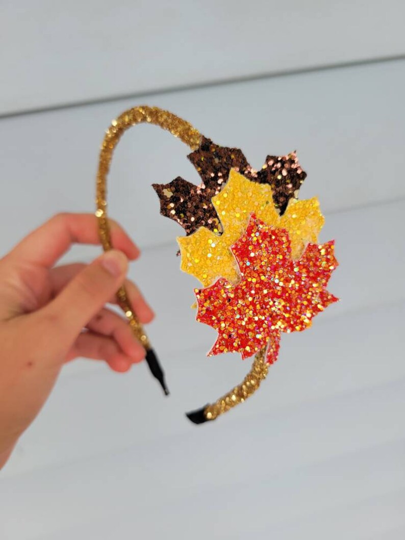 May include: A gold glitter headband with a cluster of glittery maple leaves in shades of brown, yellow, and red. The headband is held by a hand against a light background. The leaves are layered to create a 3D effect.