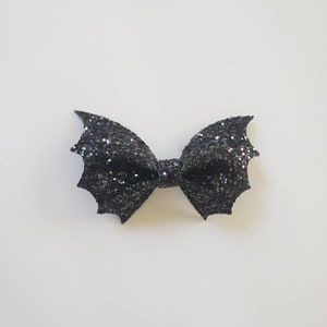 May include: A black hair bow with a glittery finish, shaped like bat wings. The accessory features a central knot and two symmetrical, pointed wing-like sides covered in dense, sparkling black glitter that reflects light across the surface.