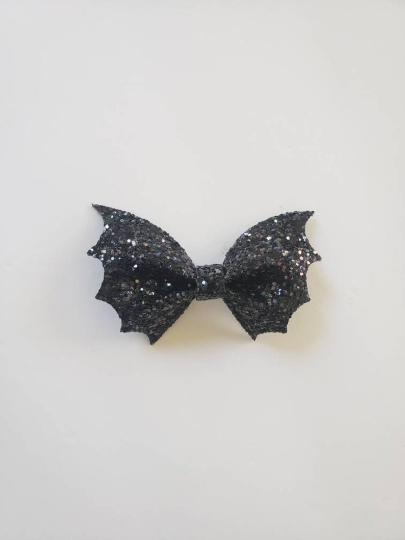 May include: A black hair bow with a glittery finish, shaped like bat wings. The accessory features a central knot and two symmetrical, pointed wing-like sides covered in dense, sparkling black glitter that reflects light across the surface.