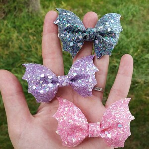 May include: Three bat-shaped hair bows featuring a heavy glitter finish. The top bow is dark grey, the middle bow is light purple, and the bottom bow is light pink. Each bow has a central knot and pointed wing-like edges with reflective sequins.