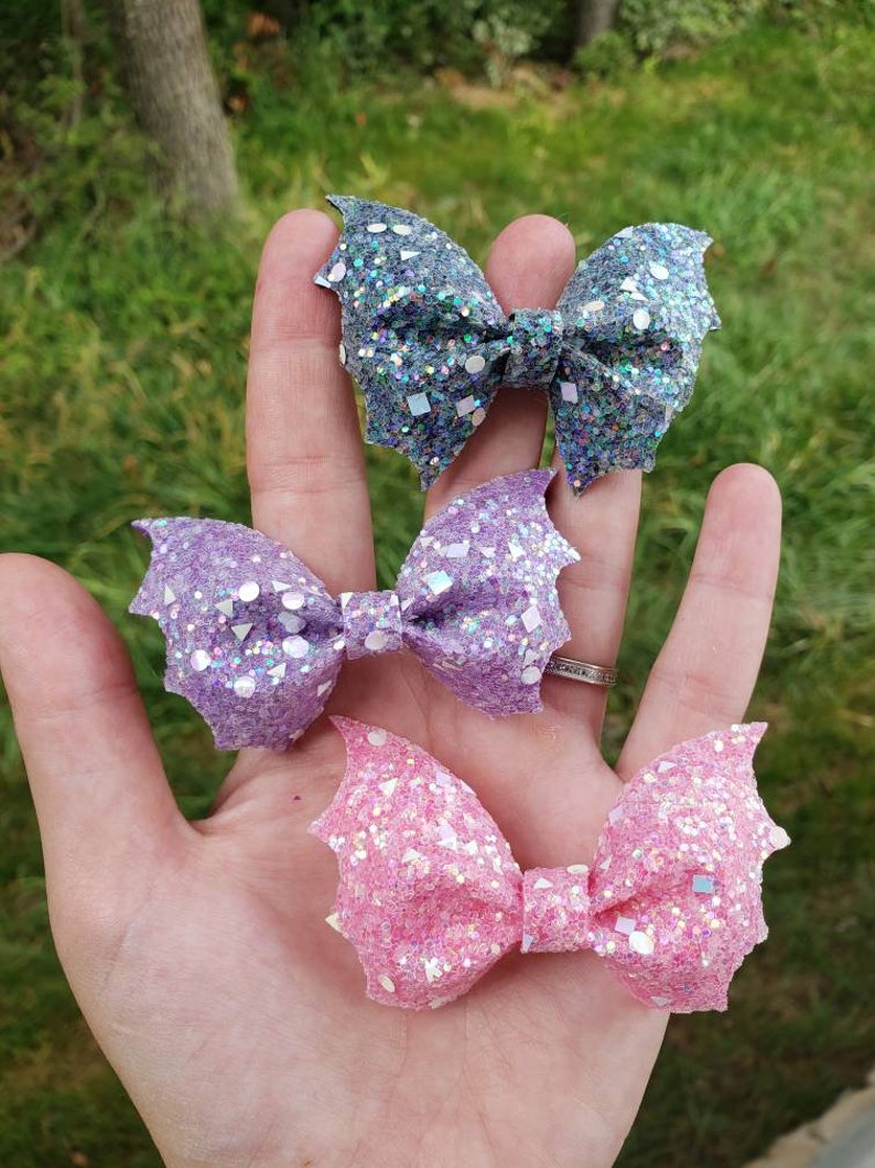 May include: Three bat-shaped hair bows featuring a heavy glitter finish. The top bow is dark grey, the middle bow is light purple, and the bottom bow is light pink. Each bow has a central knot and pointed wing-like edges with reflective sequins.
