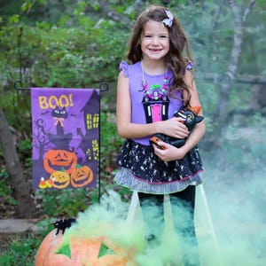 May include: A purple sleeveless top with a ruffled shoulder and a black skirt featuring a white skeleton print. The top displays a graphic of a haunted house with a pink door. A black cat figurine is held, and a purple garden flag reads BOO!.