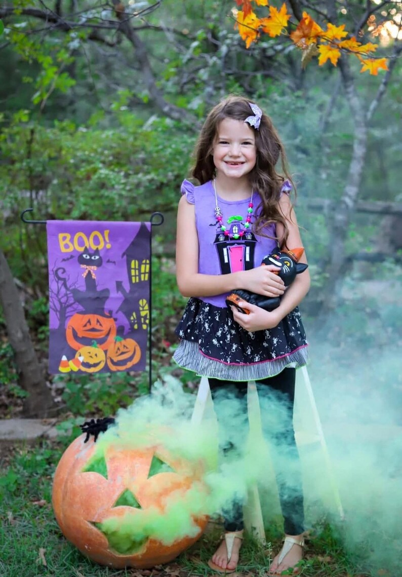 May include: A purple sleeveless top with a ruffled shoulder and a black skirt featuring a white skeleton print. The top displays a graphic of a haunted house with a pink door. A black cat figurine is held, and a purple garden flag reads BOO!.
