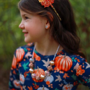 May include: A young person wearing a fall-themed headband with a glittery leaf design in shades of red, orange, and brown. They are also wearing a necklace with large, round beads and a navy blue top with a pumpkin pattern.
