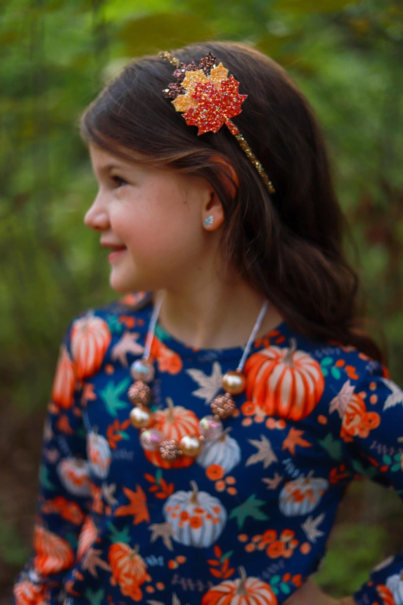 May include: A young person wearing a fall-themed headband with a glittery leaf design in shades of red, orange, and brown. They are also wearing a necklace with large, round beads and a navy blue top with a pumpkin pattern.