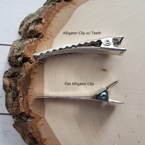 Puede incluir: Dos pinzas de cocodrilo plateadas sobre una superficie de madera. La pinza superior tiene dientes y la pinza inferior es plana. El texto "Alligator Clip w/ Teeth" est&aacute; sobre la pinza superior y el texto "Flat Alligator Clip" est&aacute; sobre la pinza inferior.