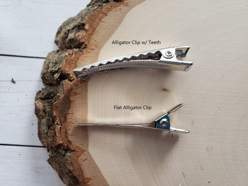 Puede incluir: Dos pinzas de cocodrilo plateadas sobre una superficie de madera. La pinza superior tiene dientes y la pinza inferior es plana. El texto "Alligator Clip w/ Teeth" est&aacute; sobre la pinza superior y el texto "Flat Alligator Clip" est&aacute; sobre la pinza inferior.