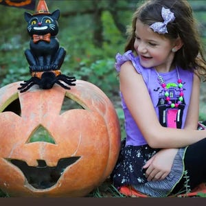 May include: A large orange carved pumpkin with a black cat figurine wearing an orange hat and bow tie perched on top, accompanied by a black spider. The cat has yellow eyes and a white grin, sitting on a textured base atop the pumpkin.
