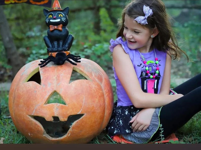 May include: A large orange carved pumpkin with a black cat figurine wearing an orange hat and bow tie perched on top, accompanied by a black spider. The cat has yellow eyes and a white grin, sitting on a textured base atop the pumpkin.