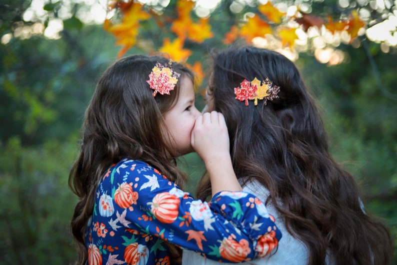 May include: Two young girls with long brown hair wearing leaf-shaped hair clips. One girl is whispering into the other's ear. The girls are wearing fall-themed clothing, with a blurred background of autumn foliage.