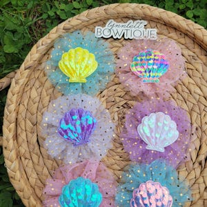 May include: A collection of six seashell-shaped hair accessories in various colors, including yellow, pink, blue, and purple. Each shell is adorned with iridescent sequins and set on a tulle flower with gold polka dots. The accessories are arranged on a woven basket. The text "lynnlett BOWTIQUE" is visible.