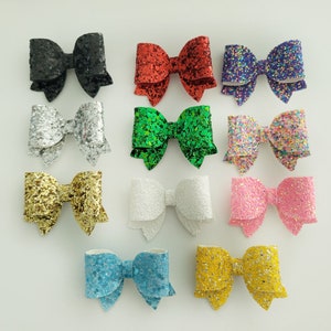 May include: Twelve glitter hair bows in various colors, including black, red, silver, green, white, pink, blue, and yellow. The bows are all approximately the same size and shape, and they are arranged in a grid pattern on a white background.