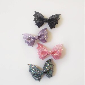 May include: Four hair bows shaped like bat wings, each covered in chunky glitter. The colors from top to bottom are black, light purple, light pink, and a dark grey with iridescent flecks. Each bow features a central knot detail.