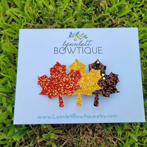 May include: A white card with the text "lynnlett BOWTIQUE" and "www.lynnlettbowtique.etsy.com" features three glittery leaf-shaped hair clips in shades of red, gold, and brown, set against a backdrop of green grass.