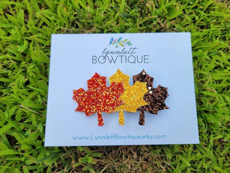 May include: A white card with the text "lynnlett BOWTIQUE" and "www.lynnlettbowtique.etsy.com" features three glittery leaf-shaped hair clips in shades of red, gold, and brown, set against a backdrop of green grass.