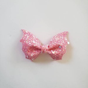 May include: A pink bow accessory featuring a dense layer of iridescent glitter and larger reflective sequins. The bow has a structured, folded shape with pointed, wing-like edges on both sides and a small central knot holding the layers together.