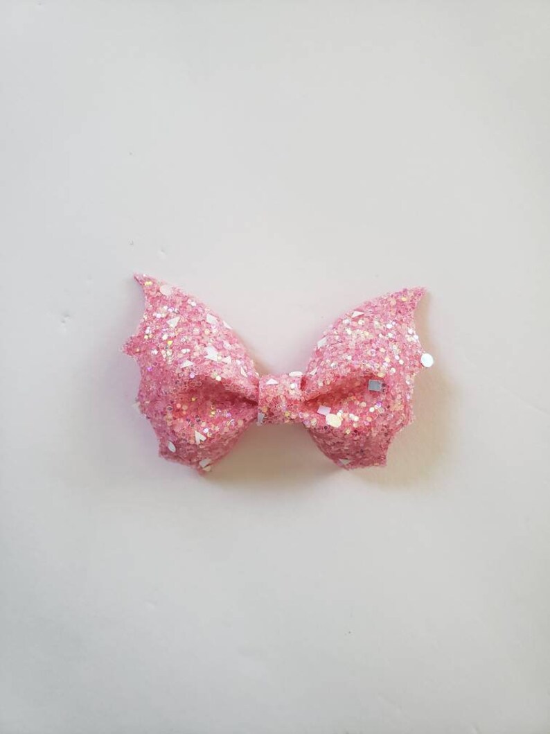 May include: A pink bow accessory featuring a dense layer of iridescent glitter and larger reflective sequins. The bow has a structured, folded shape with pointed, wing-like edges on both sides and a small central knot holding the layers together.
