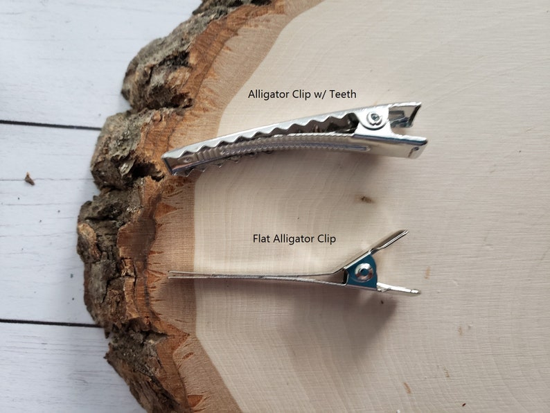 May include: Two silver-toned metal hair clips. The top clip is an alligator clip with teeth, and the bottom clip is a flat alligator clip. The text "Alligator Clip w/ Teeth" is above the top clip, and "Flat Alligator Clip" is above the bottom clip.