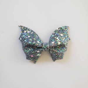 May include: A bow accessory featuring a dark grey base covered in dense, multi-colored iridescent glitter. The surface is embellished with scattered, larger reflective sequins in various geometric shapes, including circles, triangles, and squares.