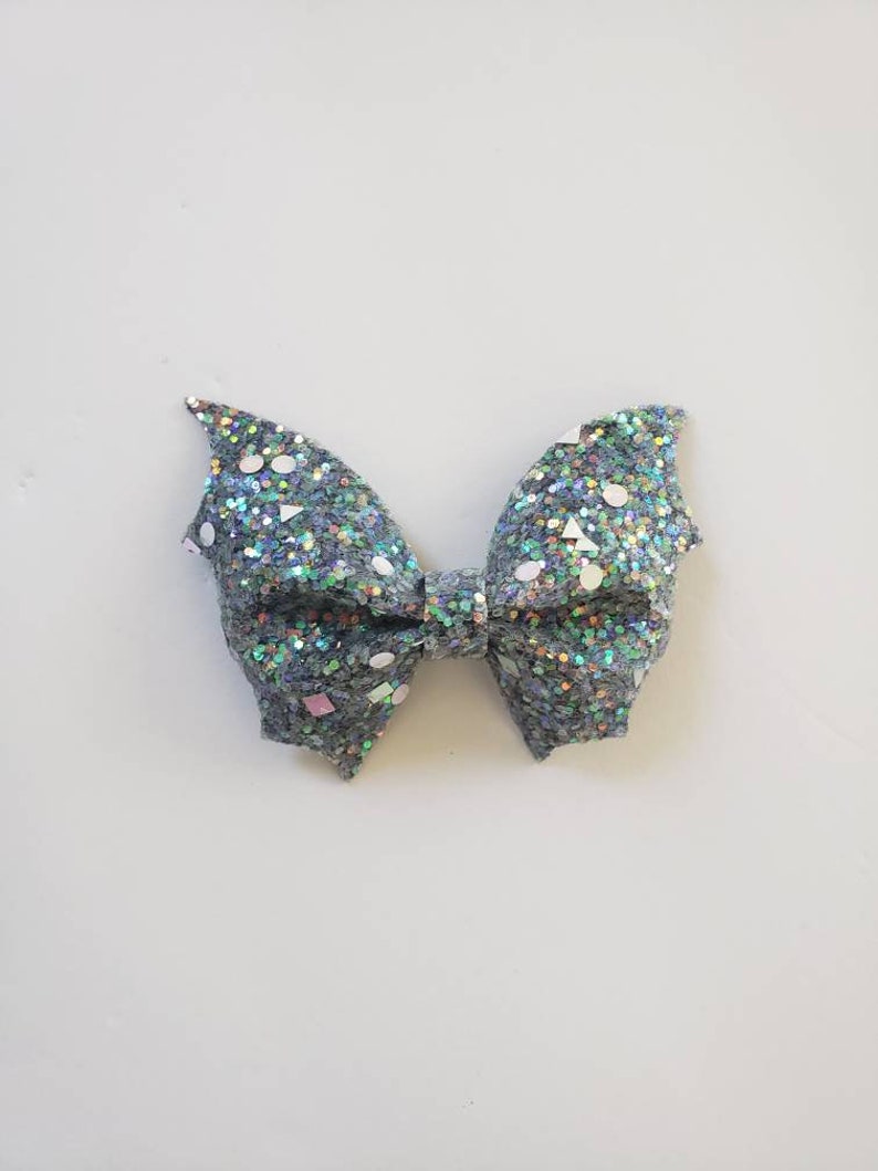 May include: A bow accessory featuring a dark grey base covered in dense, multi-colored iridescent glitter. The surface is embellished with scattered, larger reflective sequins in various geometric shapes, including circles, triangles, and squares.