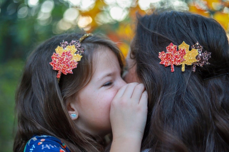 May include: A young girl and an adult with matching fall-themed hair accessories. The girl wears a headband with a glittery red, yellow, and orange leaf. The adult has a similar leaf-shaped hair clip. The background is blurred.