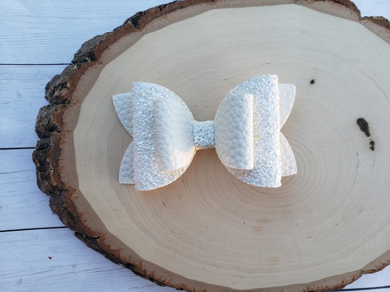 Puede incluir: Un lazo para el pelo blanco brillante con una gran forma de lazo en capas. El lazo est&aacute; sobre una superficie de madera marr&oacute;n claro.