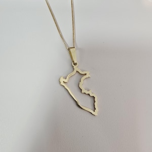 Puede incluir: Un collar con colgante dorado que representa el contorno de un país. El colgante cuelga de una fina cadena dorada. El collar se muestra sobre un fondo blanco.