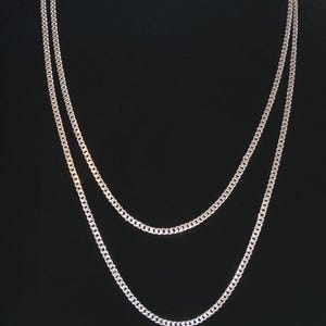 May include: Two silver-colored curb chain necklaces displayed against a black background. The necklaces are of different lengths, with one longer than the other. The chains have a shiny, metallic appearance and are likely made of metal.