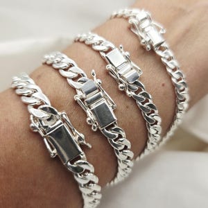 Könnte beinhalten: Vier silberfarbene Panzerkettenarmbänder mit rechteckigen Verschlüssen. Die Armbänder werden an einem Handgelenk präsentiert und zeigen das polierte Metall und die aufwendigen Kettenglieder. Klassischer Schmuck.