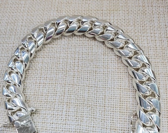 May include: A silver curb chain bracelet with a polished finish. The bracelet features a series of interlocking, rounded links and a secure clasp. The jewelry is displayed on a neutral, textured surface.