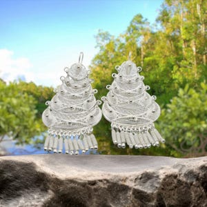 May include: A pair of silver filigree earrings. Each earring features a tiered design with intricate detailing and dangling beaded fringe. The earrings are displayed against a natural background of greenery and sky.