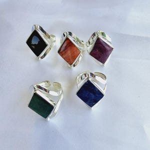 May include: Five adjustable silver rings with square gemstones in various colors: black, orange, purple, green, and blue. The rings are displayed on a white surface, showcasing the different gemstone hues and the silver band design.