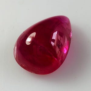 Op de afbeelding: Een traanvormige edelsteen met een diepe, levendige magenta-rode kleur en een glad, gepolijst oppervlak. De steen heeft een hoogglanzende afwerking die licht reflecteert, waardoor heldere witte accenten op het gebogen, doorschijnende lichaam ontstaan.