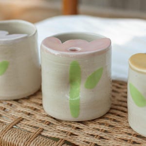 May include: Three ceramic cups with a floral design on the rim. Each cup is a different color: lavender, pink, and yellow. The cups have a light green bamboo-like design on the front.